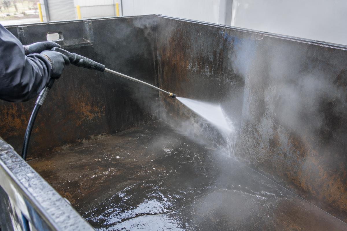 Pressure washer spraying the grimy interior of a commercial dumpster, water and steam visible